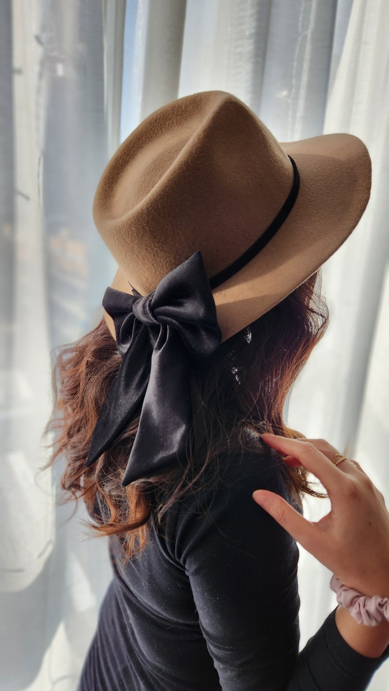 May include: A brown felt fedora hat with a black velvet ribbon and a large black satin bow. The hat is worn by a person with long brown hair.