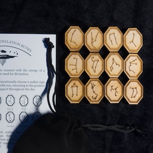 Może przedstawiać: Zestaw 12 drewnianych run z konstelacjami zodiaku w czarnym aksamitnym woreczku. Runy są sześciokątne i przedstawiają inną konstelację zodiaku na każdej stronie. Runy są dołączone do karty z instrukcjami, jak ich używać.