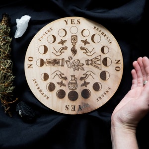 May include: A wooden circular divination board with a moon phase design. The board has the words "YES", "NO", "REPHRASE", and "UNSURE" printed around the edge. There are eight moon phases depicted in the center of the board.