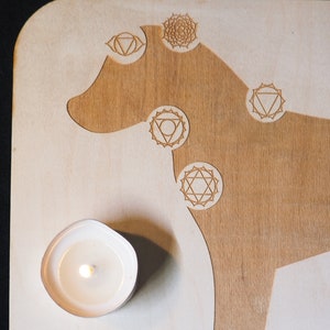 May include: A wooden board with a dog silhouette engraved on it. The dog's body is outlined with seven chakra symbols, each with a different geometric design. A small white candle is burning in the lower left corner of the board.