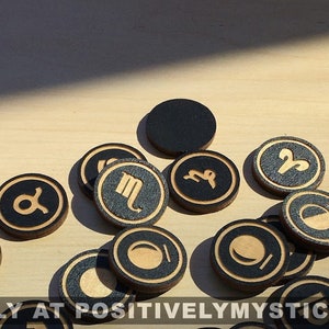 May include: A set of 15 black and gold wooden tokens featuring astrological symbols and moon phases. The tokens are scattered on a light brown wooden surface.