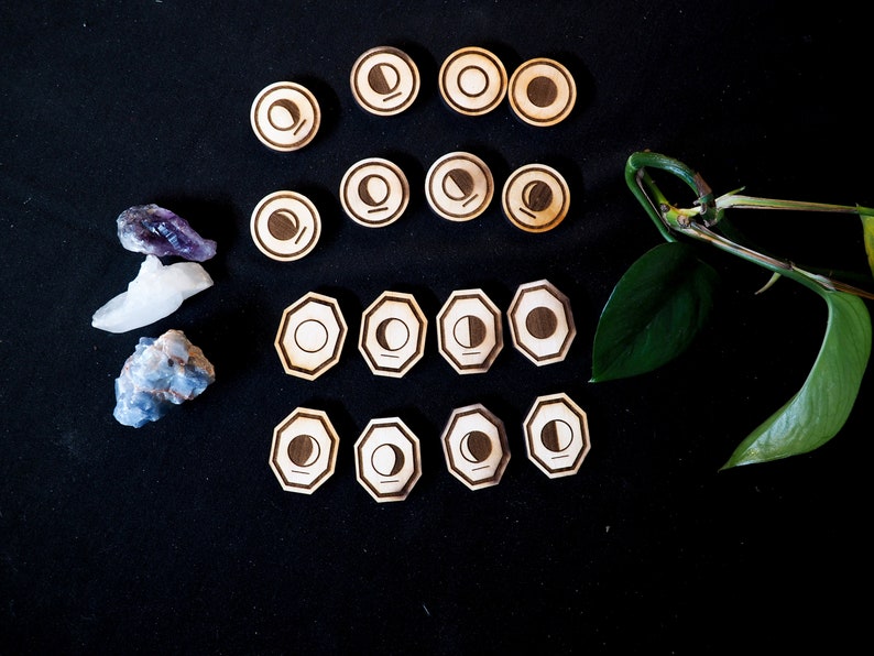 Op de afbeelding: Een set van 15 houten maanfase-tokens, met verschillende maanfasen, gerangschikt in een rasterpatroon op een zwart oppervlak. De tokens zijn in verschillende vormen en maten, sommige zijn rond en andere zijn achthoekig. Er zijn ook drie kleine kristallen aan de linkerkant van de afbeelding.