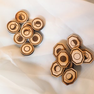 Op de afbeelding: Twee stapels houten tokens met een halve maan ontwerp. De tokens zijn rond en hebben een bruine houten afwerking. De tokens zijn gestapeld op een witte stoffen achtergrond.