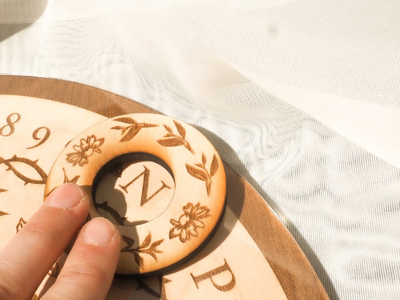 May include: A wooden circular game piece with a floral design and the letter "N" in the center. The piece is being held by a finger.