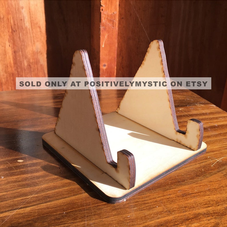 May include: A wooden display stand with a triangular shape, made of light brown wood with a darker brown border. The stand is designed to hold a small object, such as a phone or a book.  SOLD ONLY AT POSITIVELYMYSTIC ON ETSY