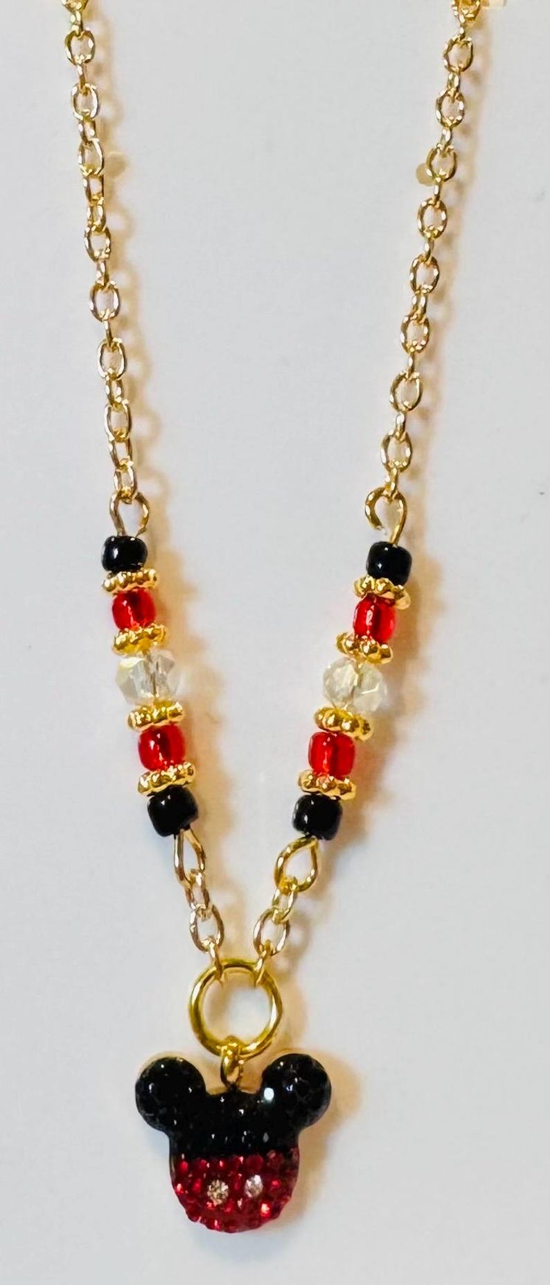 Puede incluir: Un collar de cadena dorada con un colgante de Mickey Mouse. La cadena presenta cuentas negras, rojas y transparentes. El colgante es una cabeza de Mickey Mouse negra y roja con detalles de cristal.