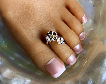 May include: Silver toe ring with two flower designs. The larger flower has a clear rhinestone in the center. The ring is on a person's toe.