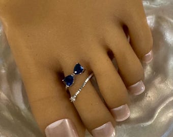 May include: A silver toe ring with a bow design featuring two blue teardrop-shaped stones. A second silver toe ring with a small clear stone is also visible. The toenails are painted with a light pink and white French manicure.