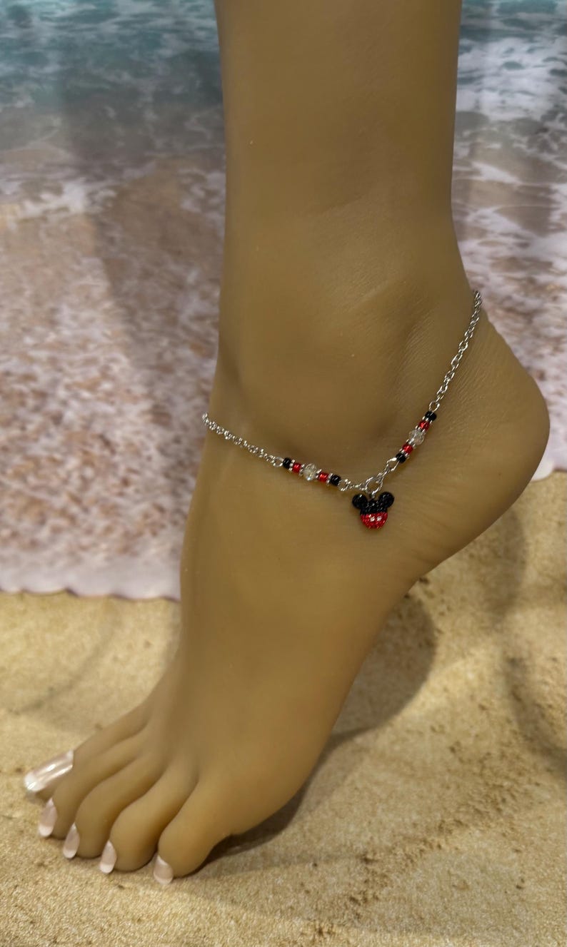 Pode incluir: Tornozeleira prateada com um pingente do Mickey Mouse. A tornozeleira apresenta uma corrente com contas vermelhas, pretas e transparentes. O pingente do Mickey Mouse &eacute; vermelho e preto. A tornozeleira &eacute; usada em um tornozelo.