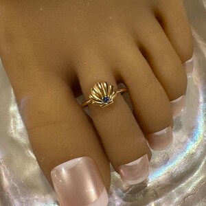 May include: A gold-colored toe ring in the shape of a seashell, with a small blue gemstone at its center. The ring is worn on a toe with a light pink and white French manicure. The ring is a piece of jewelry.