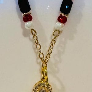 Puede incluir: Collar dorado con un colgante con una piedra transparente y brillante rodeada de cristales m&aacute;s peque&ntilde;os. La cadena tiene cuentas rojas, negras y blancas. Un accesorio encantador.