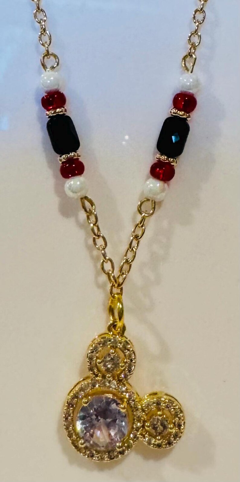 Puede incluir: Collar dorado con un colgante con una piedra transparente y brillante rodeada de cristales m&aacute;s peque&ntilde;os. La cadena tiene cuentas rojas, negras y blancas. Un accesorio encantador.