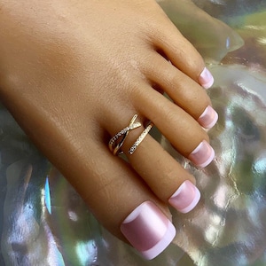 May include: Gold toe ring with clear stones, featuring a criss-cross design. The ring is worn on a foot with pink and white nail polish.