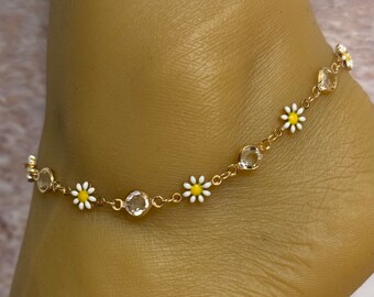 May include: Gold-toned anklet featuring alternating clear, round gemstones and small daisy flower charms with white petals and yellow centers. The anklet is worn on a foot, showcasing its delicate design and floral accents.
