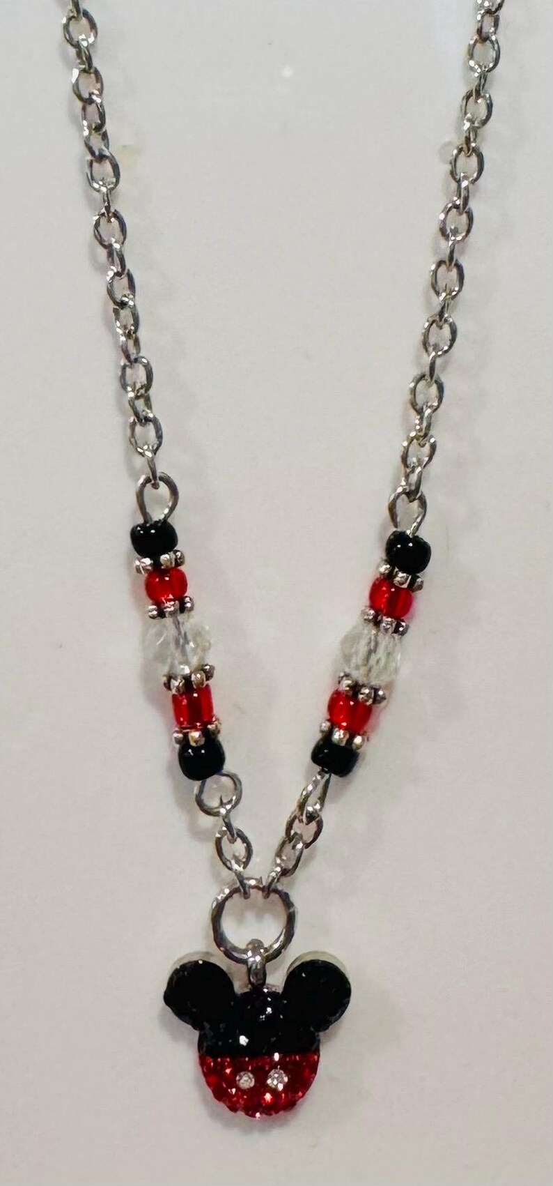 Pode incluir: Colar de corrente prateada com um pingente do Mickey Mouse. O pingente &eacute; preto e vermelho com detalhes de cristal. A corrente tem contas pretas, vermelhas e transparentes.