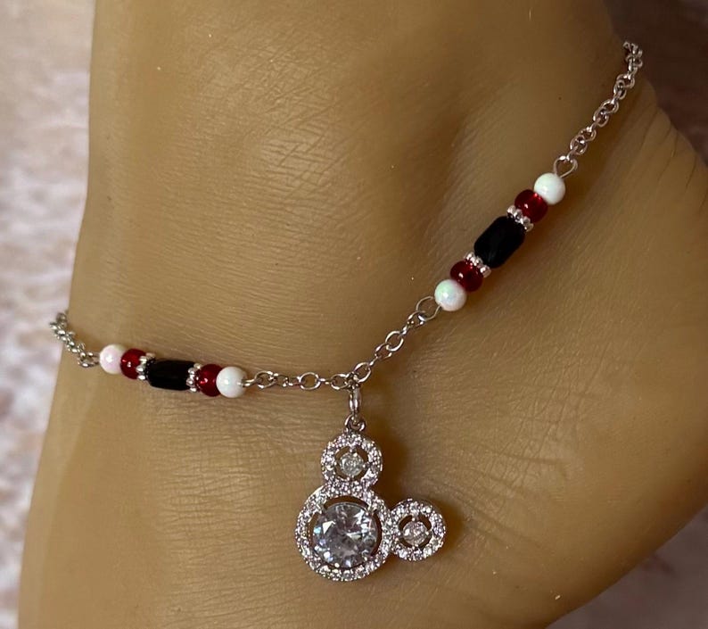 Puede incluir: Tobillera plateada con un dije de Mickey Mouse con una piedra central grande y transparente y piedras m&aacute;s peque&ntilde;as alrededor. La cadena est&aacute; adornada con cuentas rojas, negras y blancas. Un accesorio divertido.