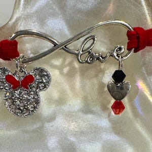 May include: A red suede bracelet with a silver infinity symbol charm that says "Love". The bracelet has two charms, one is a silver Minnie Mouse head with red bow and the other is a silver heart with a black bead and a red crystal.