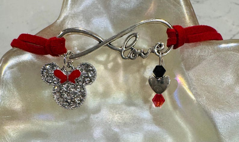 May include: A red suede bracelet with a silver infinity symbol charm that says "Love". The bracelet has two charms, one is a silver Minnie Mouse head with red bow and the other is a silver heart with a black bead and a red crystal.