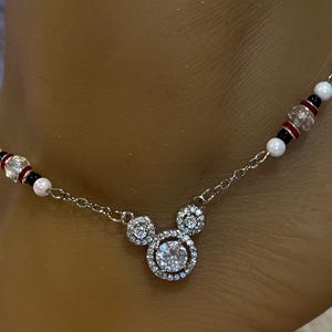 Puede incluir: Una tobillera plateada con un dise&ntilde;o inspirado en Mickey Mouse. El dije central es una piedra redonda y transparente rodeada de piedras transparentes m&aacute;s peque&ntilde;as. La cadena est&aacute; adornada con cuentas blancas, negras y rojas. Una joya delicada y encantadora.