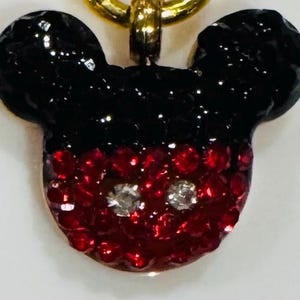 Puede incluir: Un colgante de Mickey Mouse con una cadena dorada. El colgante tiene forma de la cabeza de Mickey, con orejas negras brillantes y una cara roja brillante. Dos gemas plateadas representan los ojos.