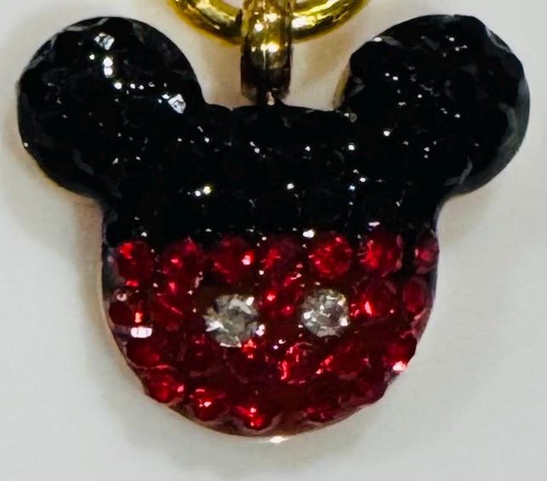 Puede incluir: Un colgante de Mickey Mouse con una cadena dorada. El colgante tiene forma de la cabeza de Mickey, con orejas negras brillantes y una cara roja brillante. Dos gemas plateadas representan los ojos.