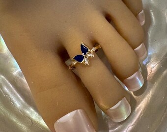 May include: A gold-toned toe ring featuring a butterfly design with navy blue enamel wings and small clear accent stones. The ring is worn on a foot with a French manicure.
