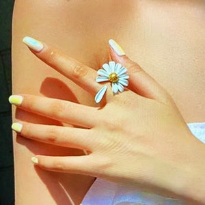 May include: A white daisy ring with a gold center. The ring is on a finger with yellow and blue nail polish.