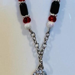 Puede incluir: Collar plateado con un colgante con tres piedras redondas claras y brillantes. La cadena est&aacute; adornada con cuentas rojas, negras y blancas. Una joya elegante.