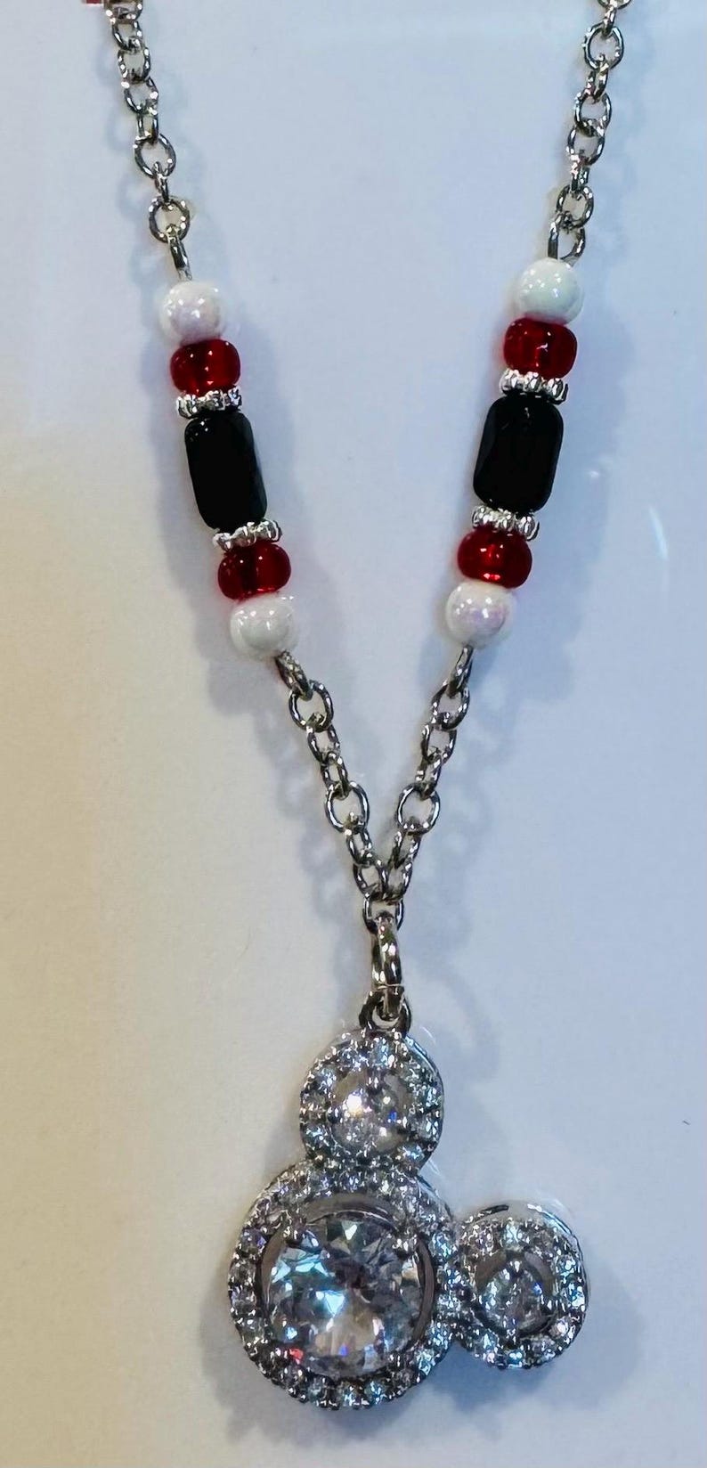 Puede incluir: Collar plateado con un colgante con tres piedras redondas claras y brillantes. La cadena est&aacute; adornada con cuentas rojas, negras y blancas. Una joya elegante.