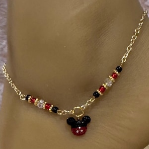 Puede incluir: Tobillera dorada con un dije de Mickey Mouse. La tobillera tiene un dise&ntilde;o de cadena con cuentas rojas, negras y transparentes. El dije de Mickey Mouse es negro y rojo con un dise&ntilde;o de pedrer&iacute;a.