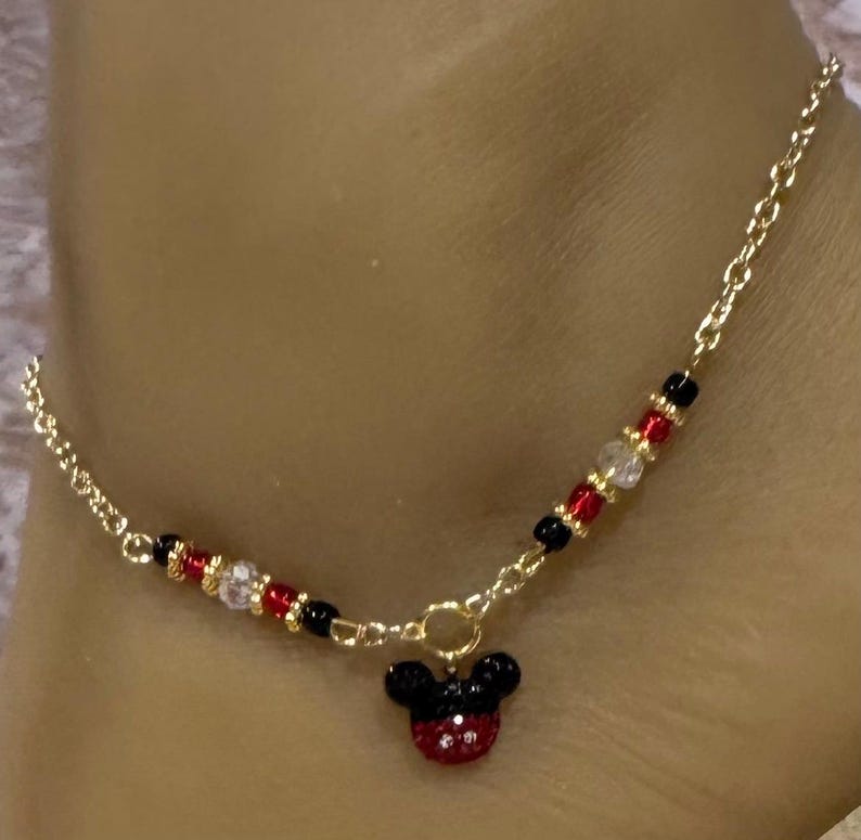 Puede incluir: Tobillera dorada con un dije de Mickey Mouse. La tobillera tiene un dise&ntilde;o de cadena con cuentas rojas, negras y transparentes. El dije de Mickey Mouse es negro y rojo con un dise&ntilde;o de pedrer&iacute;a.