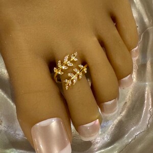 May include: A gold-colored toe ring featuring a delicate leaf design adorned with small, clear gemstones. The ring is worn on a toe with a light pink and white nail polish.