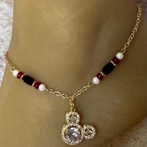 Puede incluir: Tobillera dorada con un colgante de Mickey Mouse con una piedra central grande y transparente y piedras m&aacute;s peque&ntilde;as. La cadena est&aacute; adornada con cuentas rojas, negras y blancas. Una joya delicada y encantadora.