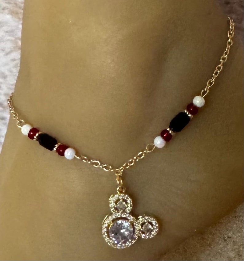 Puede incluir: Tobillera dorada con un colgante de Mickey Mouse con una piedra central grande y transparente y piedras m&aacute;s peque&ntilde;as. La cadena est&aacute; adornada con cuentas rojas, negras y blancas. Una joya delicada y encantadora.