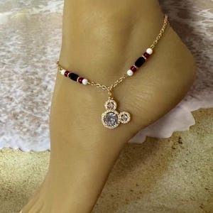 Puede incluir: Tobillera dorada con un dije de Mickey Mouse con una piedra transparente en el centro. La cadena est&aacute; adornada con cuentas rojas, negras y blancas. La tobillera se muestra en un pie, con un fondo de playa.