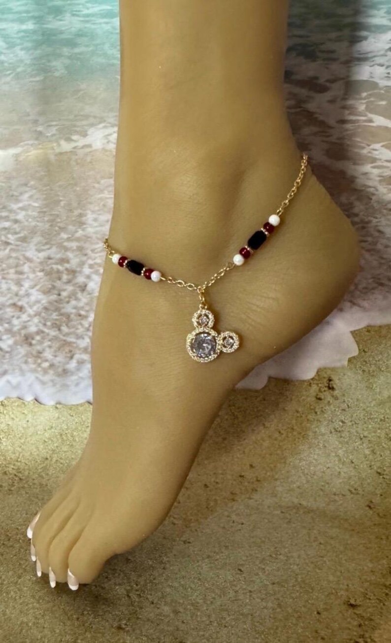 Puede incluir: Tobillera dorada con un dije de Mickey Mouse con una piedra transparente en el centro. La cadena est&aacute; adornada con cuentas rojas, negras y blancas. La tobillera se muestra en un pie, con un fondo de playa.