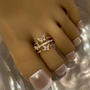 May include: A gold-toned toe ring featuring a layered design with butterfly accents and clear gemstones. The ring is worn on a foot with a light skin tone and light pink nail polish.