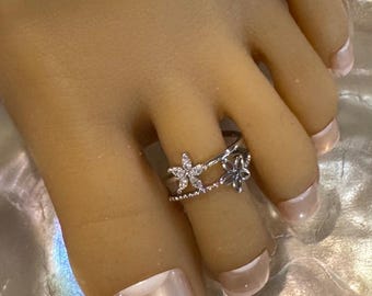 May include: A silver-toned toe ring featuring two flower designs. One ring has a row of small beads, and the other has a single flower. The ring is worn on a foot with a light pink nail polish.