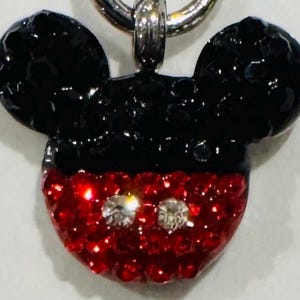 Pode incluir: Um pingente do Mickey Mouse com uma corrente prateada. As orelhas s&atilde;o pretas e o rosto vermelho com duas pedras preciosas claras e brilhantes para os olhos. O pingente &eacute; provavelmente feito de cristal ou strass.