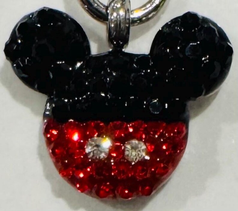 Pode incluir: Um pingente do Mickey Mouse com uma corrente prateada. As orelhas s&atilde;o pretas e o rosto vermelho com duas pedras preciosas claras e brilhantes para os olhos. O pingente &eacute; provavelmente feito de cristal ou strass.