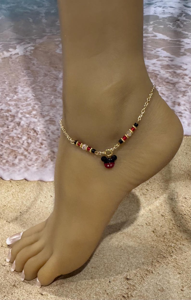 Puede incluir: Tobillera dorada con un dije de Mickey Mouse. La tobillera tiene un dise&ntilde;o de cadena con cuentas rojas, negras y transparentes. El dije de Mickey Mouse es negro y rojo. En un pie, con fondo de playa.