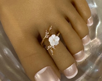 May include: A gold-toned toe ring featuring a delicate white flower and small, sparkling accents. The ring is designed with a floral motif and is worn on a foot with a French manicure.