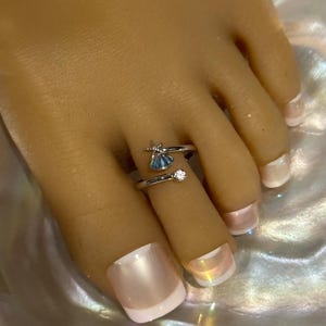 May include: A silver-toned open toe ring featuring a small starfish and a blue seashell charm on one end, with a single clear round stone set on the opposite end. The band is adjustable and wraps around the toe, showcasing a polished metallic finish.