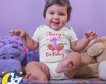 May include: A white onesie with a pink and red graphic that says "Berry 1st Birthday" with strawberries and stars. A baby is wearing the onesie and sitting on a purple and brown plush toy.