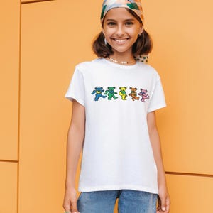 May include: A young person wearing a white t-shirt with a colorful graphic of dancing bears. The person is also wearing blue jeans and a patterned headscarf.