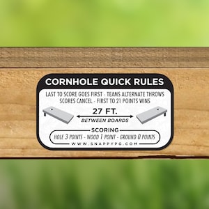 May include: White sticker with black text outlining the rules for a cornhole game. The text reads "Cornhole Quick Rules", "Last to score goes first - Teams alternate throws", "Scores cancel - First to 21 points wins", "27 ft. Between Boards", "Scoring", "Hole 3 points - Wood 1 point - Ground 0 points", and "www.snappyg.com".
