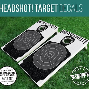 May include: Two white regulation-sized cornhole boards with black and white target decals. The decals feature a silhouette of a person with a bullseye target on the chest. The text "HEADSHOT!" is printed on each board.