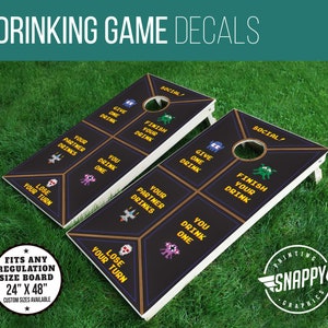 May include: Two black and white regulation size cornhole boards with custom decals. The decals feature a pixelated design with the text "Social!" and phrases like "Give One Drink", "Finish Your Drink", and "Lose Your Turn".