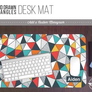 May include: A white desk mat with a geometric pattern of hand-drawn triangles in shades of orange, teal, red, and black. The mat has a grid pattern and the text "Aiden" is printed in the lower right corner.