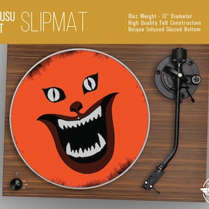 May include: Orange vinyl record slipmat with a black and white illustration of a cat's face with a wide open mouth and sharp teeth. The slipmat is 12 inches in diameter and weighs 16 ounces. It is made of high-quality felt with a unique glazed bottom.